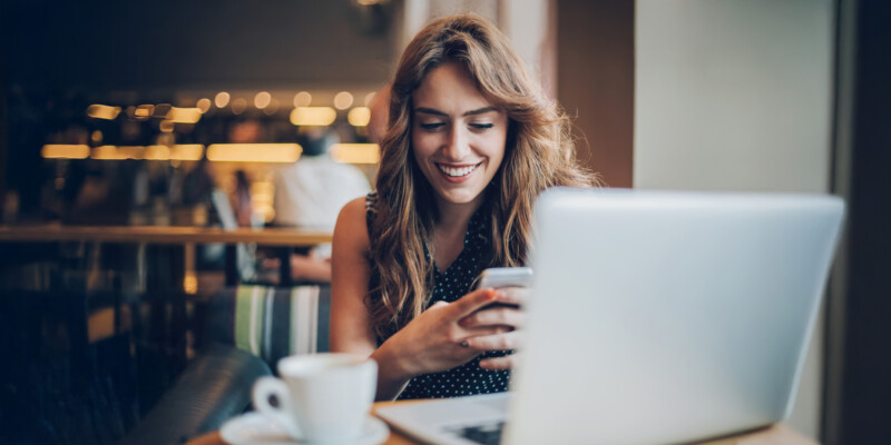 Lächelnde Frau, die ihr Smartphone an einem Cafétisch mit Laptop und Kaffeetasse benutzt.