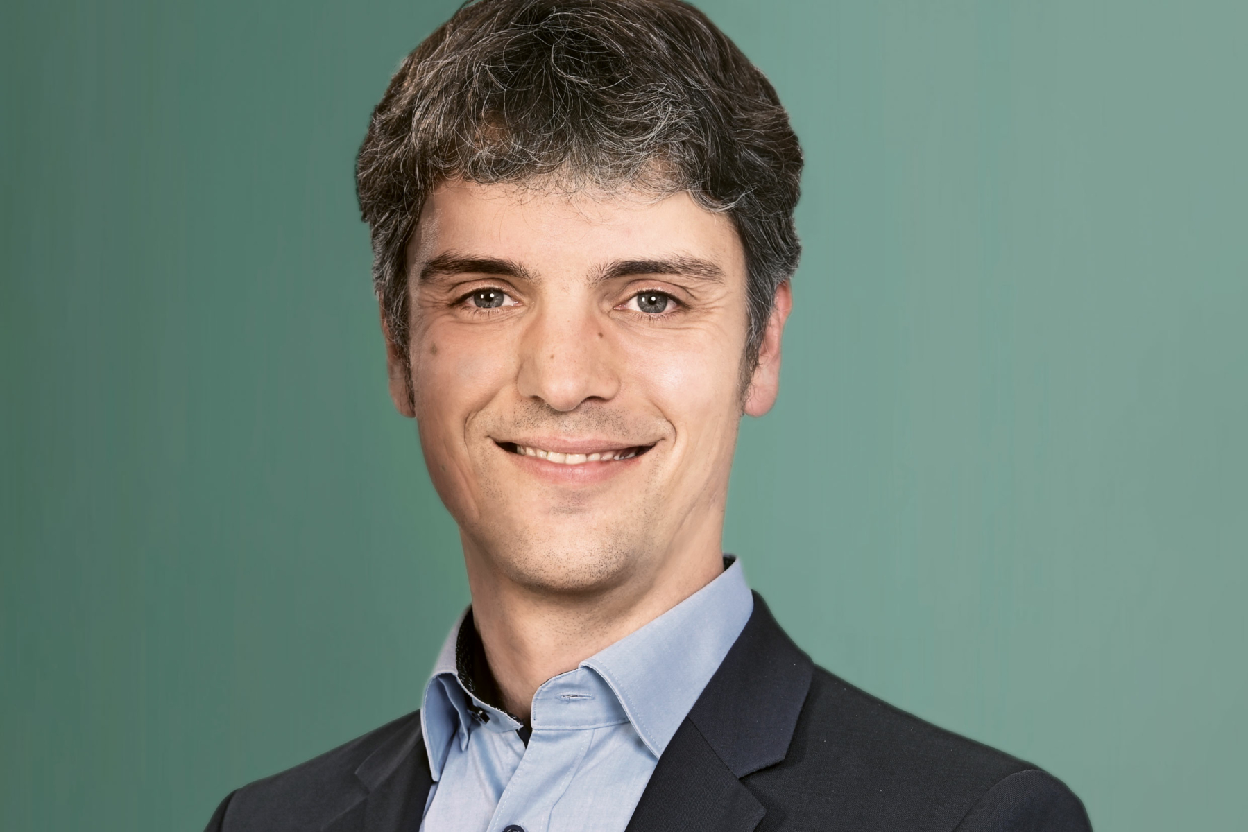 Ein Mann im Anzug lächelt vor einem schlichten grünen Hintergrund.