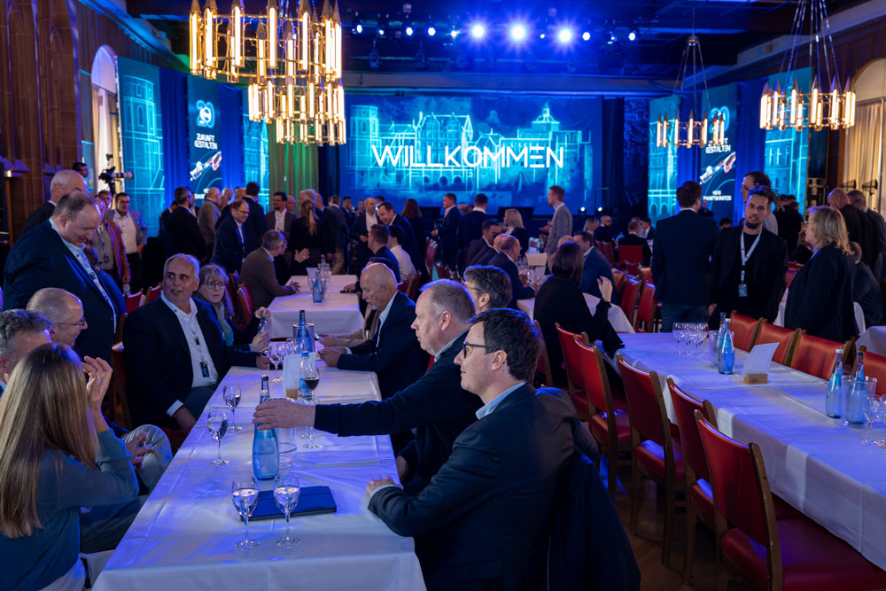 hsag-19 Die Menschen versammelten sich in einer großen Halle, in der vorne auf einer Leinwand "Willkommen" angezeigt wurde.