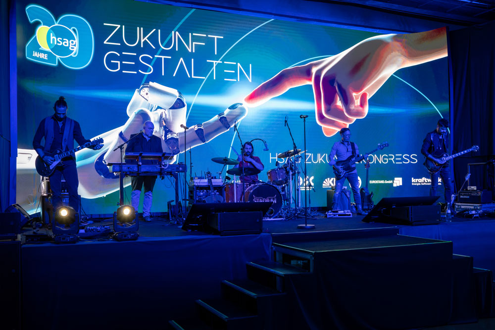 hsag-216 Eine Band tritt auf der Bühne vor einem futuristischen Bildschirm mit einer Roboterhand und deutschem Text auf.