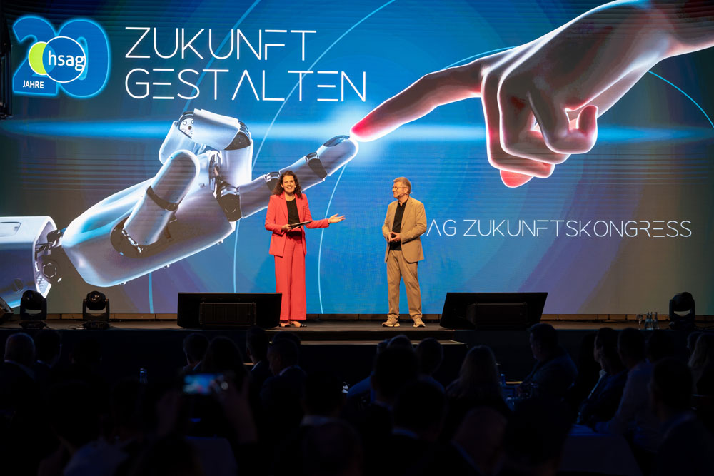 hsag-23 Zwei Sprecher auf der Bühne mit einem großen Bildschirm, der eine Roboterhand zeigt, die auf eine menschliche Hand zeigt.