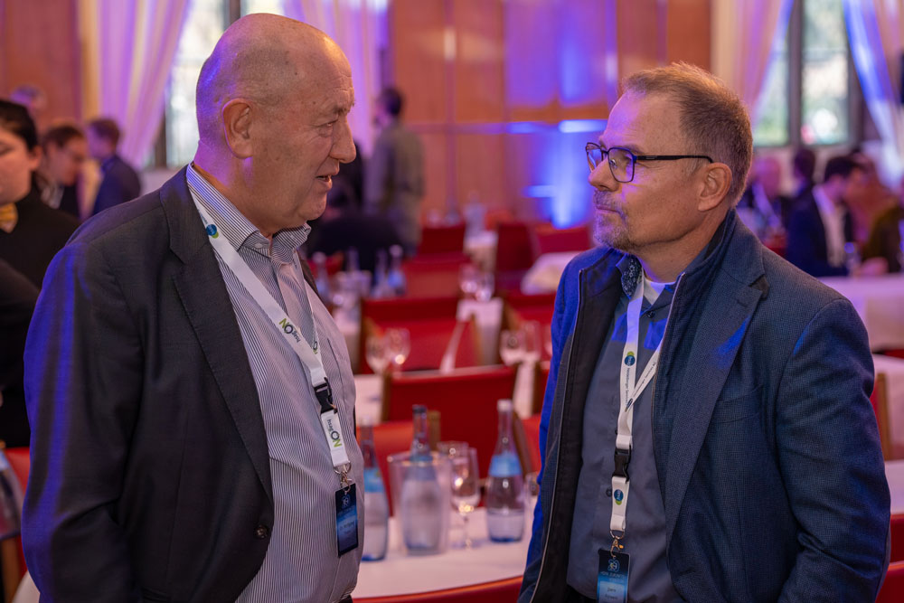 hsag-39 Zwei Männer im Geschäftsanzug unterhalten sich auf einer Konferenz mit Menschen und Tischen im Hintergrund.