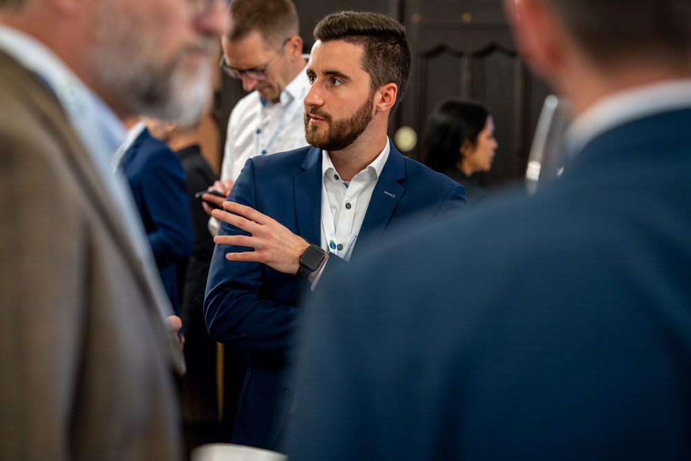 hsag-4 Ein Mann im Anzug spricht bei einem Networking-Event, andere Personen sind im Vorder- und Hintergrund unscharf.