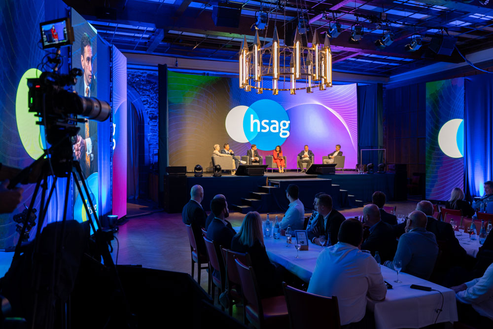 hsag-73 Podiumsdiskussion auf der Bühne mit Publikum, bunter Beleuchtung und hsag-Logo auf der Leinwand.