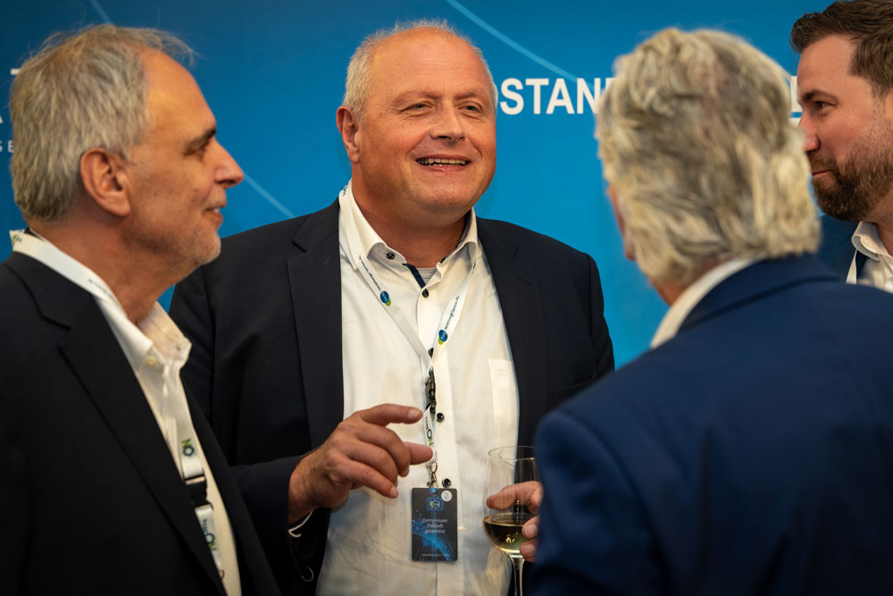 hsag-86 Vier Männer in Anzügen unterhalten sich und lächeln bei einer beruflichen Veranstaltung, einer hält ein Glas Wein in der Hand.