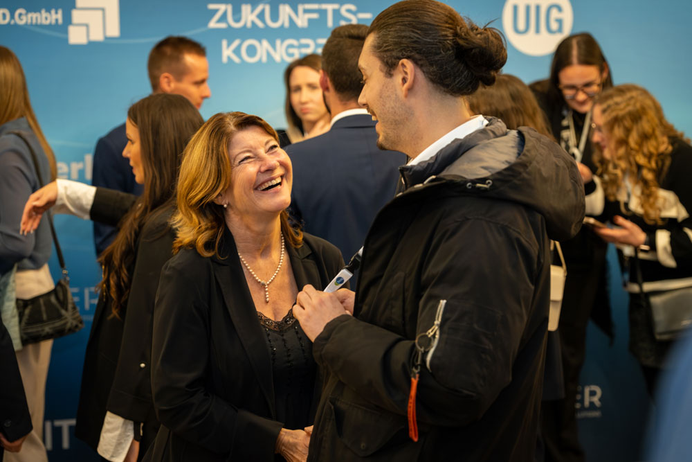 hsag-91 Zwei Personen lachen und unterhalten sich bei einer beruflichen Veranstaltung mit anderen im Hintergrund.
